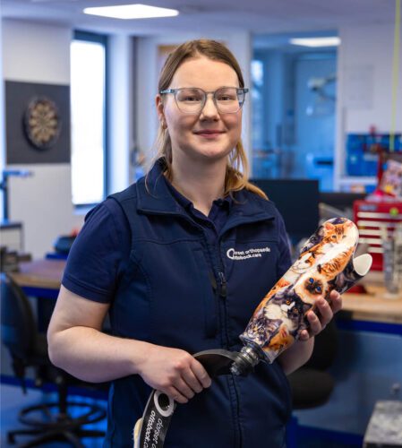 A picture of Bex stood in an office holding a prosthetic leg.