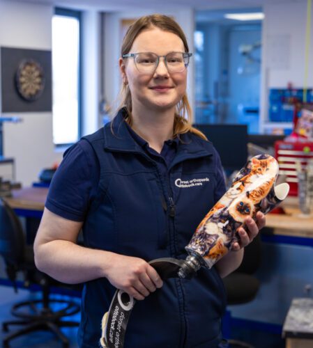 A picture of Bex stood in an office holding a prosthetic leg.