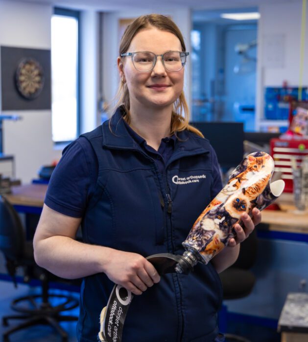 A picture of Bex stood in an office holding a prosthetic leg.
