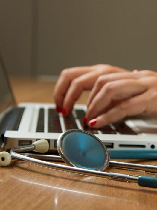 A person typing into a laptop, with a stethoscope next to them on the desk.