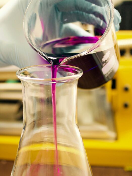 A technician wearing gloves, pouring purple dye from a beaker, into a flask.