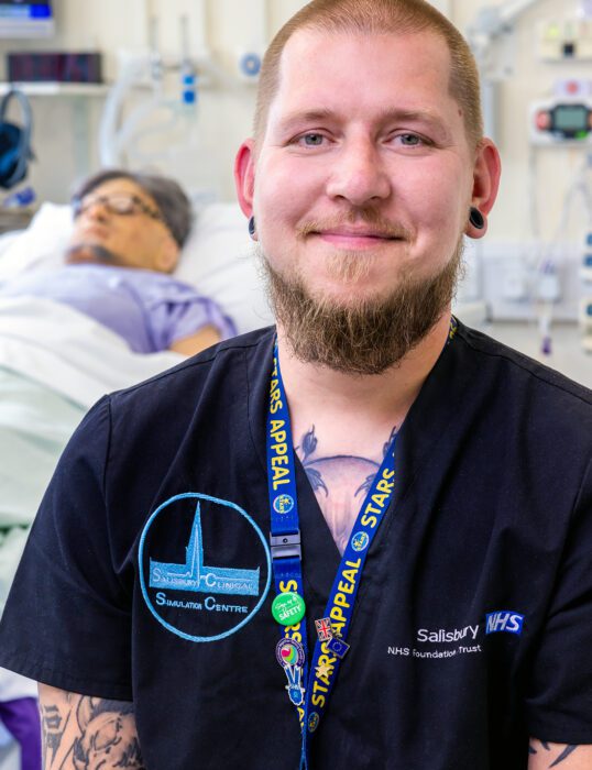 Colin Kuberka in a hospital ward, next to a simulation dummy lying in bed.