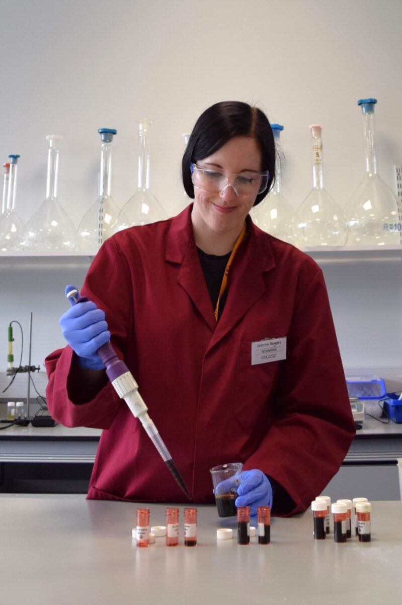 Jasmine preparing samples in the lab with a pipette, wearing a lab coat, safety glasses, and protective gloves.