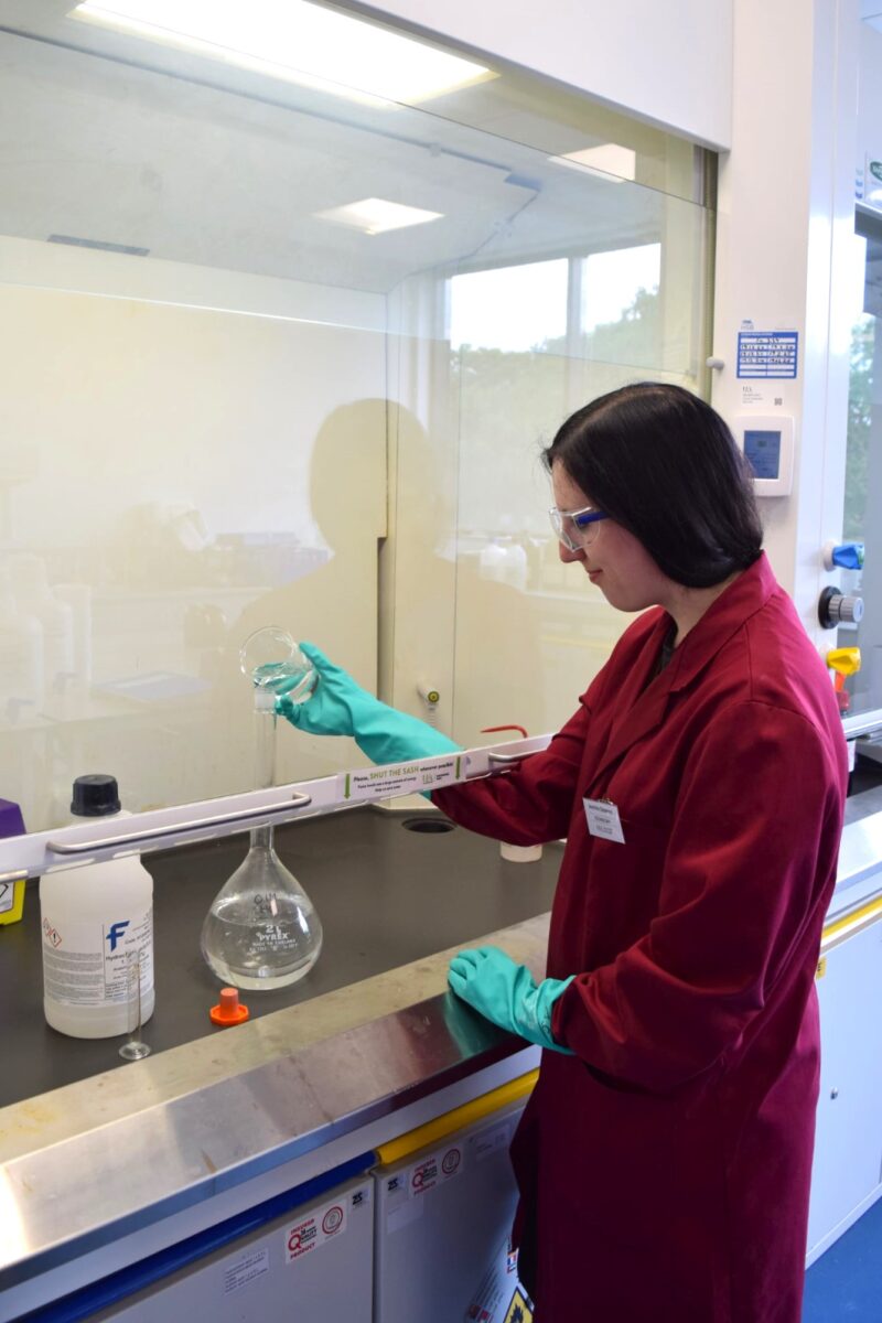 Jasmine mixing chemicals together in a fume cupboard, wearing a lab coat, safety glasses, and protective gloves.
