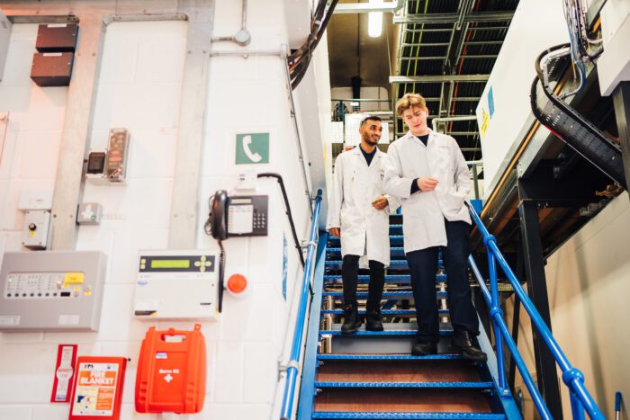 Two workers in lab coats descending stairs in factory